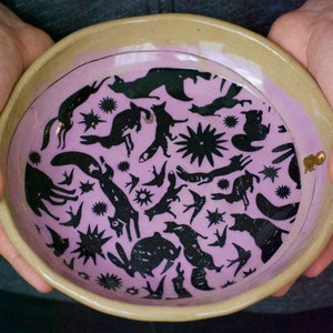 May include: A ceramic bowl with a light pink glaze and a black and white design of animals and stars. The bowl has a beige rim.