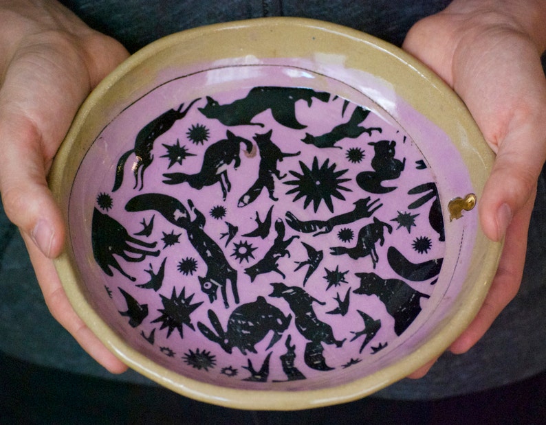 May include: A ceramic bowl with a light pink glaze and a black and white design of animals and stars. The bowl has a beige rim.