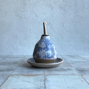 May include: A ceramic oil cruet with a pear-shaped body featuring a blue and white toile pattern. The base is unglazed tan clay, and it sits on a matching ceramic saucer. A stainless steel pour spout is inserted into the top opening.