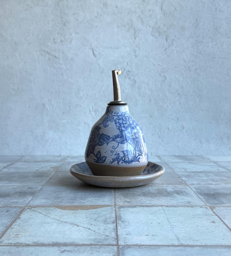 May include: A ceramic oil cruet with a pear-shaped body featuring a blue and white toile pattern. The base is unglazed tan clay, and it sits on a matching ceramic saucer. A stainless steel pour spout is inserted into the top opening.