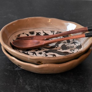 Puede incluir: Dos cuencos de cer&aacute;mica marr&oacute;n con un dise&ntilde;o en blanco y negro de p&aacute;jaros y otros animales. Los cuencos est&aacute;n apilados uno encima del otro y una cuchara y un tenedor de madera descansan sobre los cuencos.