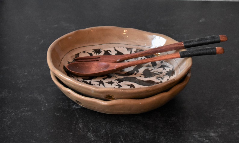 Puede incluir: Dos cuencos de cer&aacute;mica marr&oacute;n con un dise&ntilde;o en blanco y negro de p&aacute;jaros y otros animales. Los cuencos est&aacute;n apilados uno encima del otro y una cuchara y un tenedor de madera descansan sobre los cuencos.