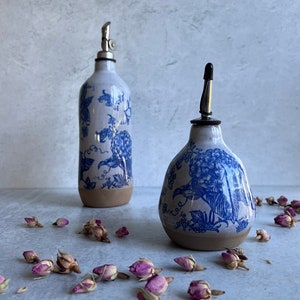 May include: Two ceramic oil dispensers with a blue and white floral pattern and a natural clay base. The taller bottle has a metal pour spout, while the shorter, wider bottle features a black cap. Dried pink rosebuds are scattered around.
