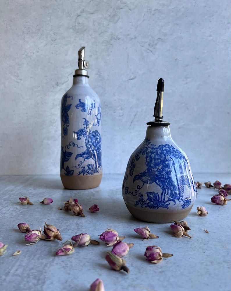 May include: Two ceramic oil dispensers with a blue and white floral pattern and a natural clay base. The taller bottle has a metal pour spout, while the shorter, wider bottle features a black cap. Dried pink rosebuds are scattered around.
