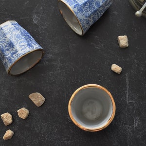 May include: Four ceramic teacups with a blue and white floral pattern and gold trim. Three of the cups are upright and one is laying on its side. There are several brown sugar cubes scattered on a black surface.
