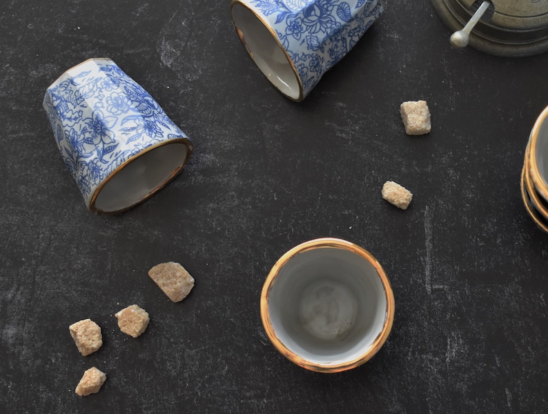 May include: Four ceramic teacups with a blue and white floral pattern and gold trim. Three of the cups are upright and one is laying on its side. There are several brown sugar cubes scattered on a black surface.