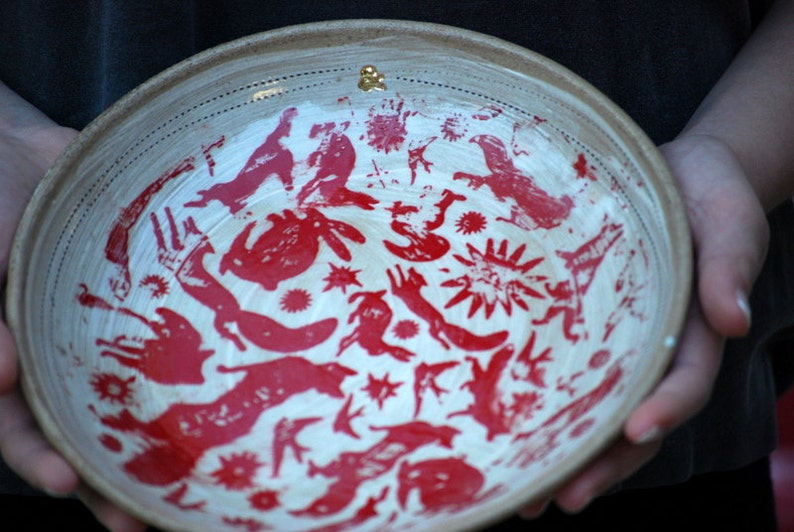 May include: A white ceramic bowl with a red and white design. The bowl is decorated with a repeating pattern of stylized animals and stars. The bowl is being held by a person's hands.