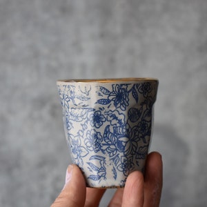 May include: A white ceramic cup with a blue floral pattern and a gold rim. The cup is being held in a hand.