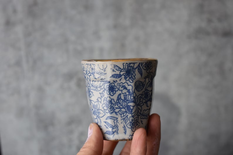 May include: A white ceramic cup with a blue floral pattern and a gold rim. The cup is being held in a hand.