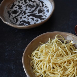 Puede incluir: Primer plano de un bol de espaguetis en un bol de cer&aacute;mica marr&oacute;n con un dise&ntilde;o en blanco y negro. El bol est&aacute; sobre una superficie negra.