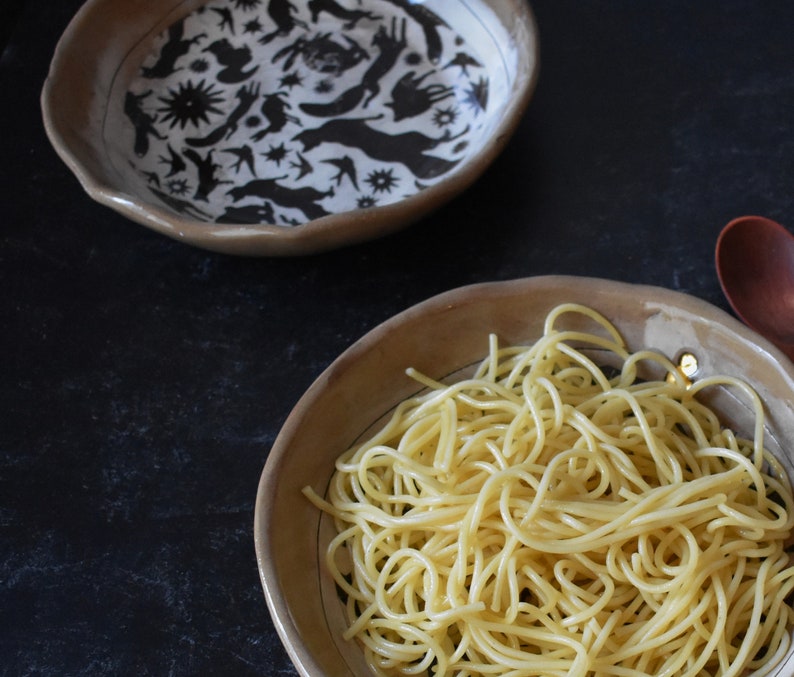 Puede incluir: Primer plano de un bol de espaguetis en un bol de cer&aacute;mica marr&oacute;n con un dise&ntilde;o en blanco y negro. El bol est&aacute; sobre una superficie negra.