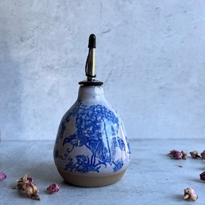 May include: A ceramic oil cruet with a rounded base and a narrow neck, featuring a blue floral pattern on a light gray background. The cruet is topped with a stainless steel pourer spout that has a small black plastic cap on the top.
