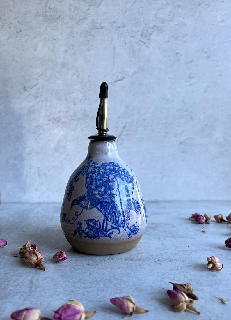 May include: A ceramic oil cruet with a rounded base and a narrow neck, featuring a blue floral pattern on a light gray background. The cruet is topped with a stainless steel pourer spout that has a small black plastic cap on the top.