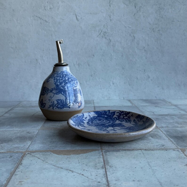 May include: A ceramic oil cruet with a blue and white floral pattern and a metal pour spout, paired with a matching small round ceramic dish featuring a blue and white floral design on a light tan base.
