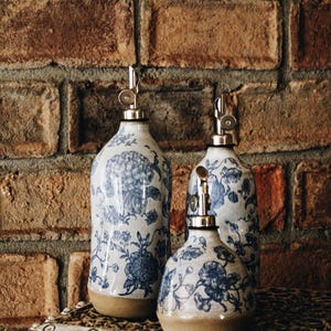 May include: Three ceramic dispensers with a blue and white floral pattern. Each bottle features a smooth, rounded body with a tan base and a silver-colored metal pour spout at the top. The set includes one tall bottle and two shorter, rounded bottles.