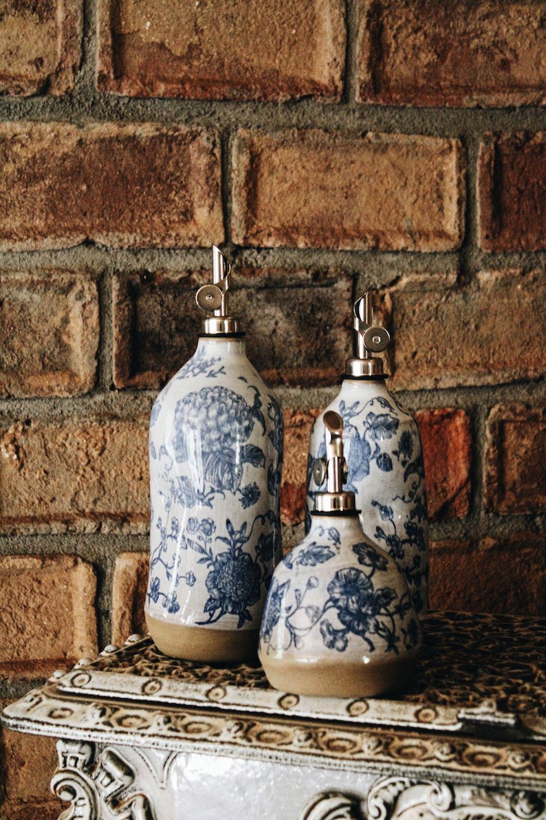 May include: Three ceramic dispensers with a blue and white floral pattern. Each bottle features a smooth, rounded body with a tan base and a silver-colored metal pour spout at the top. The set includes one tall bottle and two shorter, rounded bottles.