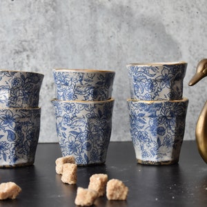 May include: Six blue and white ceramic cups with gold trim stacked in pairs. The cups have a floral pattern and are arranged on a black surface. A gold swan sugar dispenser is on the right side of the image.