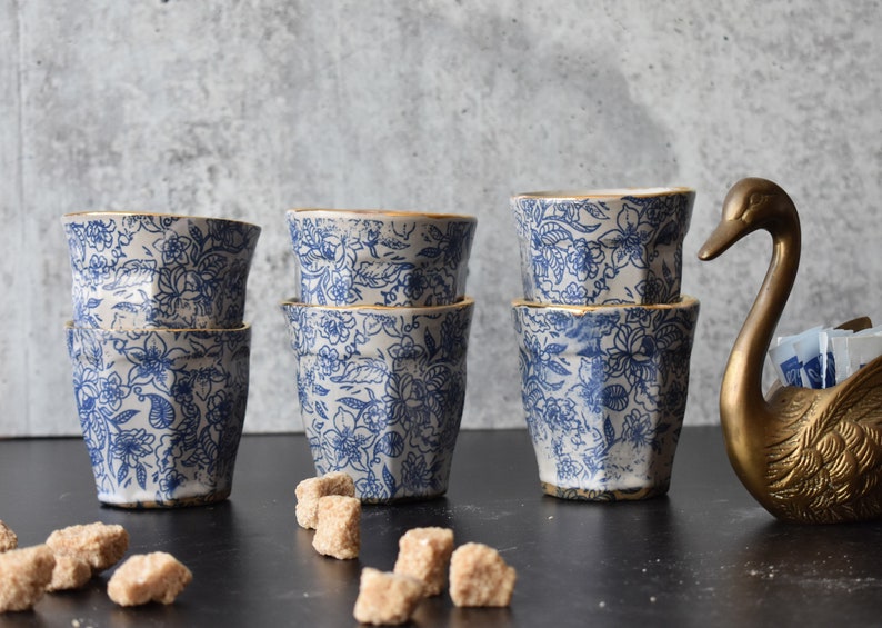 May include: Six blue and white ceramic cups with gold trim stacked in pairs. The cups have a floral pattern and are arranged on a black surface. A gold swan sugar dispenser is on the right side of the image.