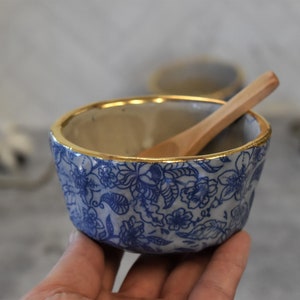 May include: A small, blue and white floral patterned ceramic bowl with a gold rim. A small wooden spoon rests inside the bowl. The bowl is likely used for serving dips, sauces, or small portions of food.