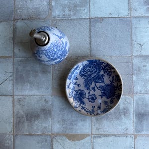 May include: A ceramic oil dispenser and a matching small plate, both featuring a blue and white floral pattern. The dispenser has a metallic pour spout, while the plate is round with a detailed botanical design in deep blue ink.