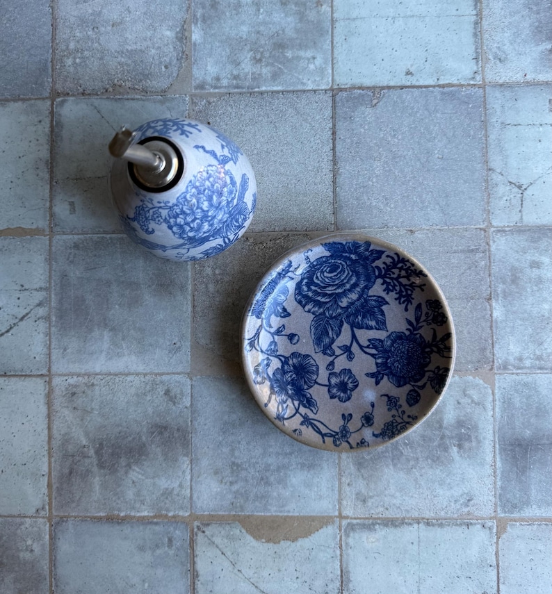 May include: A ceramic oil dispenser and a matching small plate, both featuring a blue and white floral pattern. The dispenser has a metallic pour spout, while the plate is round with a detailed botanical design in deep blue ink.