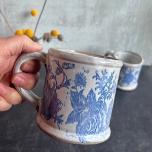 May include: A ceramic mug with a blue floral pattern. The mug has a white background with blue floral designs. The handle is white and the rim is a light brown color. A second mug is visible in the background.