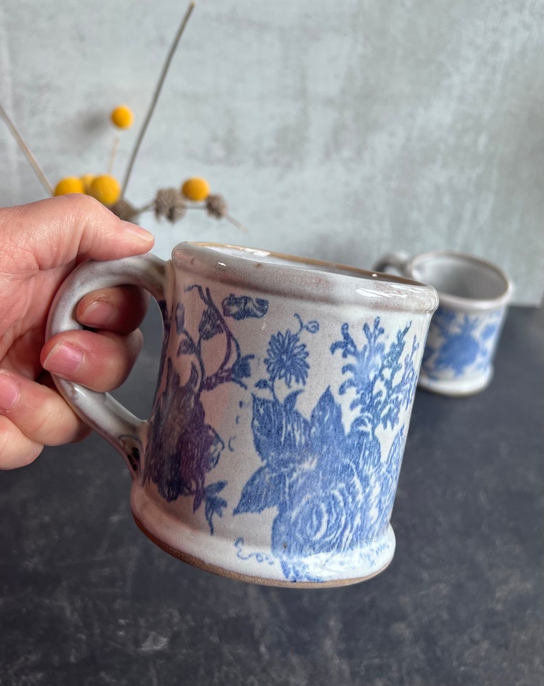 May include: A ceramic mug with a blue floral pattern. The mug has a white background with blue floral designs. The handle is white and the rim is a light brown color. A second mug is visible in the background.