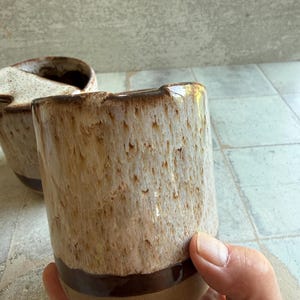 May include: Two ceramic mugs with a textured, speckled glaze in shades of beige and brown. The mugs have a dark brown band near the base and a slightly irregular rim. One mug is in focus, held by a hand, while the other is blurred in the background.