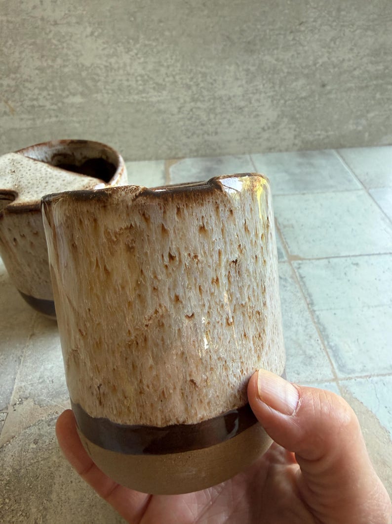 May include: Two ceramic mugs with a textured, speckled glaze in shades of beige and brown. The mugs have a dark brown band near the base and a slightly irregular rim. One mug is in focus, held by a hand, while the other is blurred in the background.