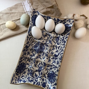 Puede incluir: Una fuente rectangular de cer&aacute;mica con un estampado floral azul contiene cinco huevos blancos. Tambi&eacute;n se ven dos huevos decorativos y una servilleta de lino beige. La fuente tiene un borde beige.