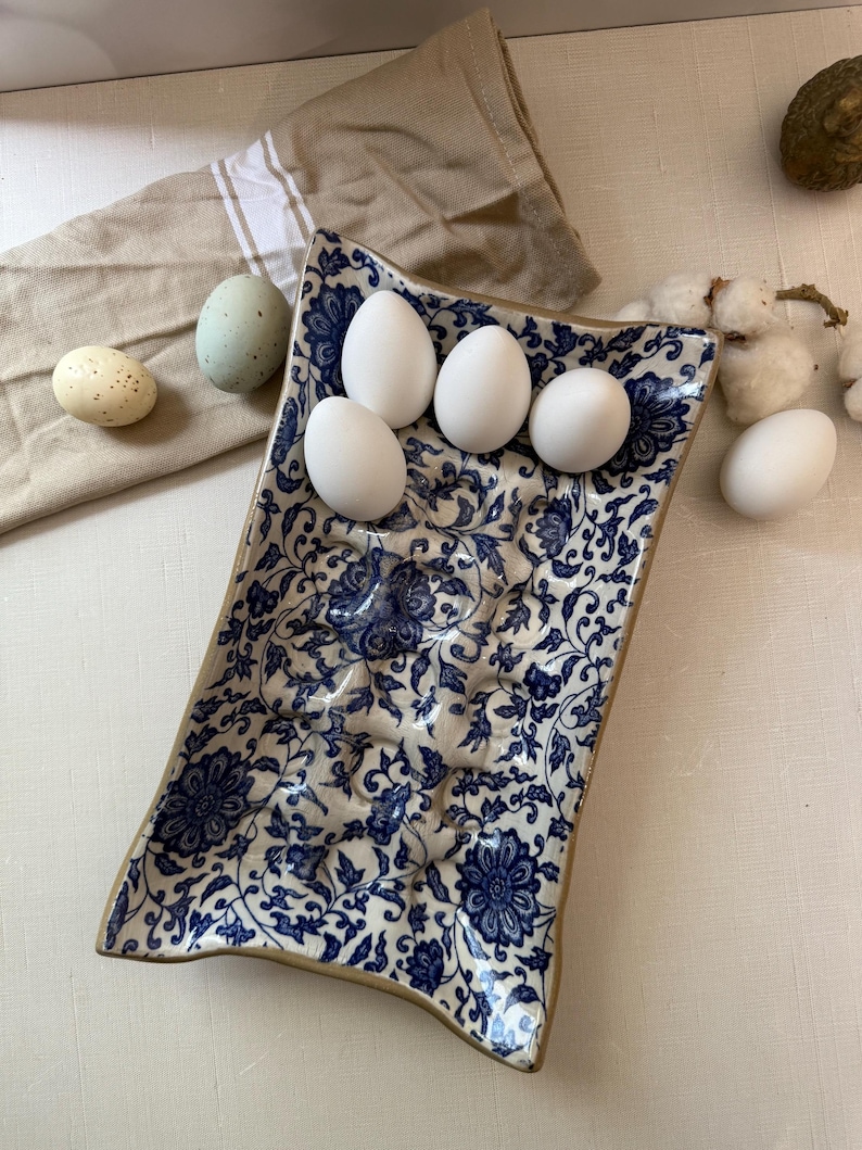 Puede incluir: Una fuente rectangular de cer&aacute;mica con un estampado floral azul contiene cinco huevos blancos. Tambi&eacute;n se ven dos huevos decorativos y una servilleta de lino beige. La fuente tiene un borde beige.