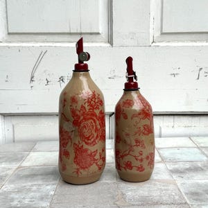 May include: Two ceramic bottles with red floral designs and red pump tops. The bottles are different sizes and are sitting on a tiled surface.