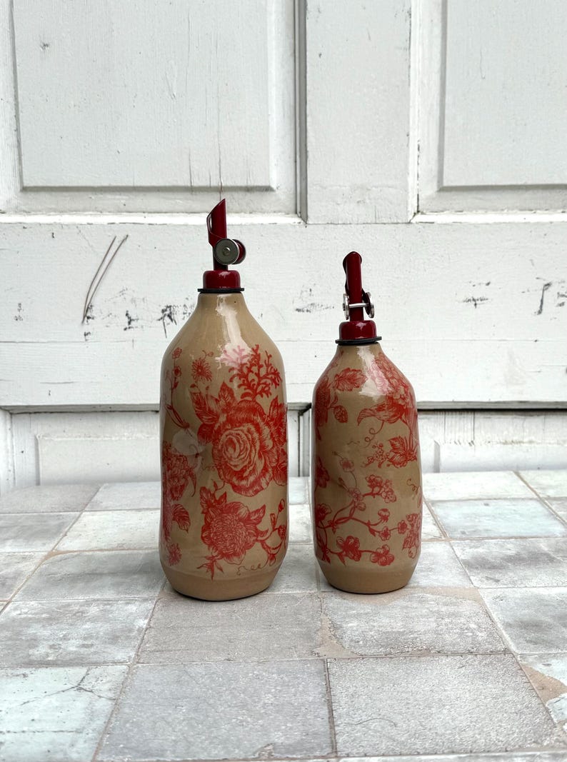 May include: Two ceramic bottles with red floral designs and red pump tops. The bottles are different sizes and are sitting on a tiled surface.