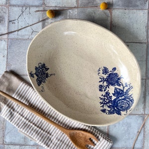 May include: A white oval ceramic serving bowl with a speckled glaze and blue floral designs. The bowl is on a striped beige and brown fabric with a wooden spoon.