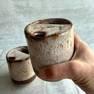 May include: Two ceramic travel mugs with a speckled beige and brown glaze. The mugs have a dark brown band near the base and a unique, asymmetrical opening at the top. One mug is held in a hand.