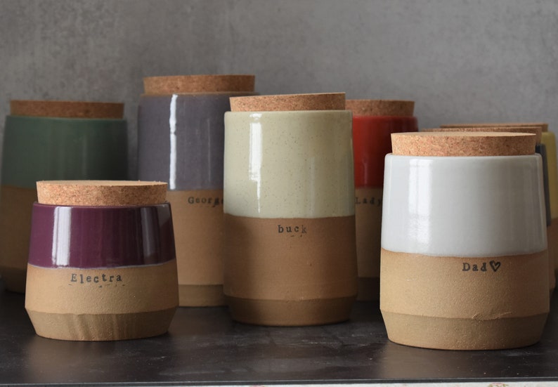 Op de afbeelding: Een verzameling keramische potten met kurken deksels. De potten zijn in verschillende kleuren, waaronder groen, paars, wit en rood, met een natuurlijke bruine basis. Elke pot heeft een naam erop gedrukt, zoals "Electra" en "Dad".