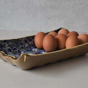 Puede incluir: Una bandeja de cer&aacute;mica para huevos con exterior beige e interior con estampado floral azul. El borde est&aacute; adornado con pintura dorada. La bandeja contiene una docena de huevos marrones.