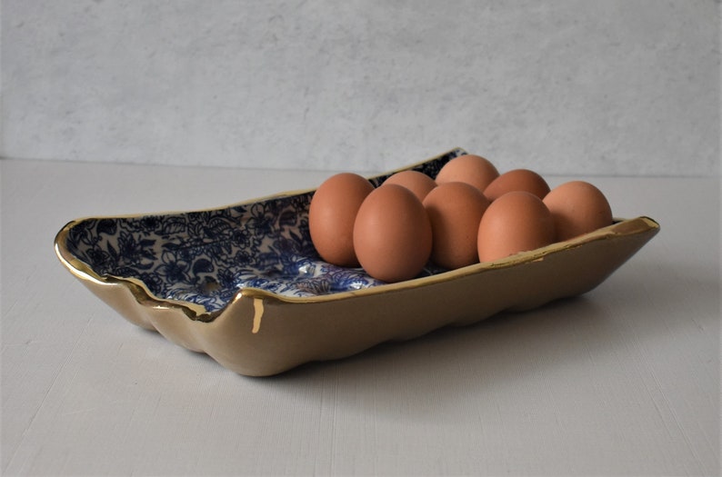 Puede incluir: Una bandeja de cer&aacute;mica para huevos con exterior beige e interior con estampado floral azul. El borde est&aacute; adornado con pintura dorada. La bandeja contiene una docena de huevos marrones.