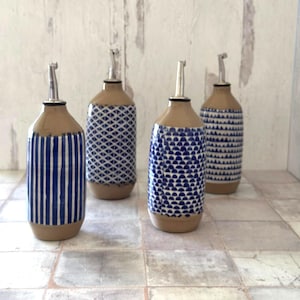 May include: Four ceramic oil bottles with stainless steel spouts. The bottles have a tan base and neck, with blue and white patterns. Designs include vertical stripes, geometric shapes, and semi-circles. The bottles are arranged on a tiled surface.