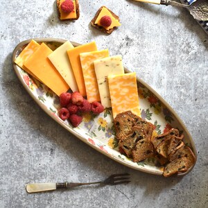 K&ouml;nnte beinhalten: Eine ovale Platte mit floralem Muster, auf der verschiedene K&auml;sesorten, Cracker und Himbeeren liegen. Die Platte befindet sich auf einem grauen Hintergrund.