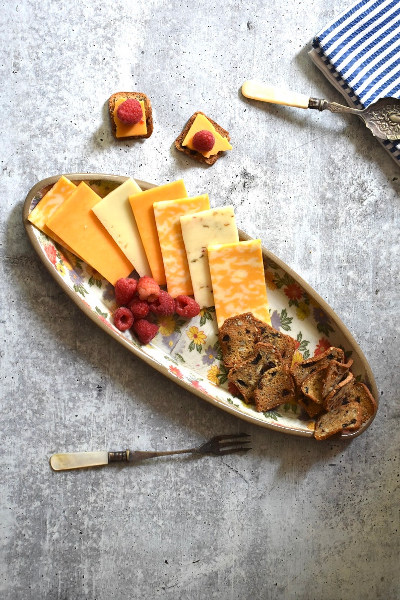 K&ouml;nnte beinhalten: Eine ovale Platte mit floralem Muster, auf der verschiedene K&auml;sesorten, Cracker und Himbeeren liegen. Die Platte befindet sich auf einem grauen Hintergrund.