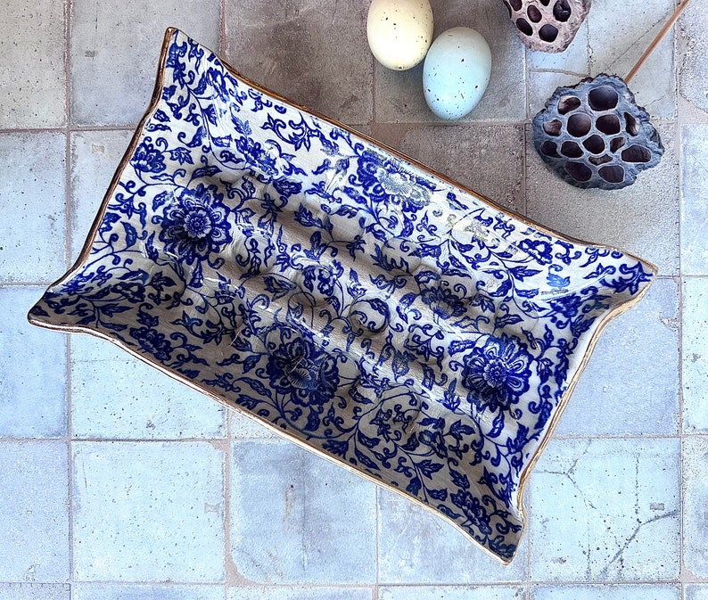 Puede incluir: Una fuente rectangular de cer&aacute;mica con un estampado floral azul y blanco y ribete dorado. La fuente tiene una forma ligeramente curvada y est&aacute; llena de un dise&ntilde;o floral detallado. El fondo incluye huevos decorativos y una vaina de loto seca.
