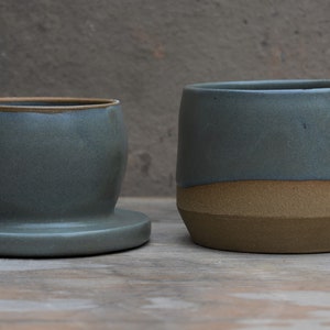 Peut inclure: Deux pots en c&eacute;ramique dans les tons de bleu-gris et de marron. Le pot de gauche a une forme arrondie avec une base. Le pot de droite est cylindrique avec un design bicolore. Les deux sont sur une surface en bois.