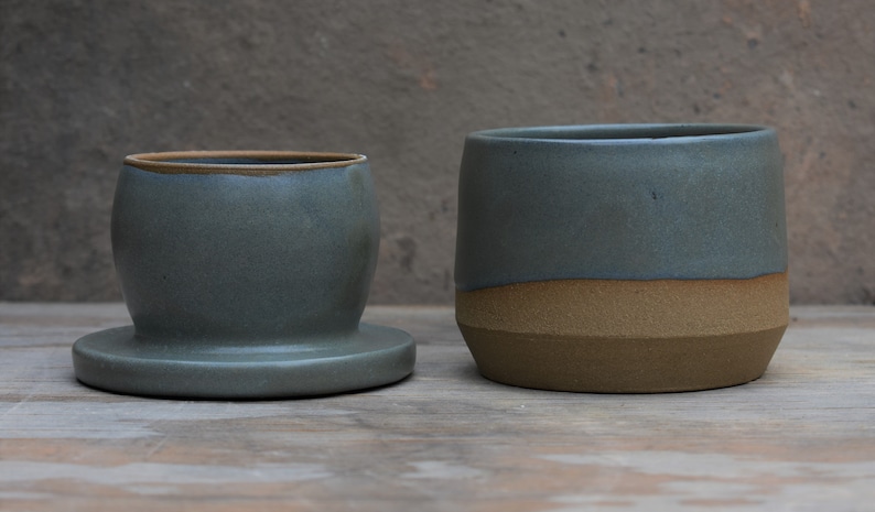 Peut inclure: Deux pots en c&eacute;ramique dans les tons de bleu-gris et de marron. Le pot de gauche a une forme arrondie avec une base. Le pot de droite est cylindrique avec un design bicolore. Les deux sont sur une surface en bois.