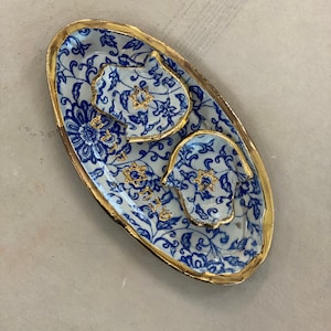 Op de afbeelding: Een ovaal keramisch serveerblad met een blauw-wit bloemmotief en gouden rand. De lade heeft twee kleinere, handvormige schalen en een kleine schaal met gouden steraccenten. De lade heeft Hebreeuwse tekst.