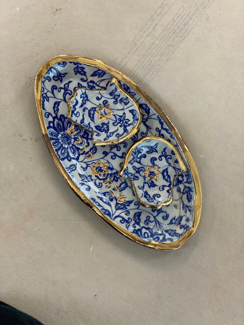Op de afbeelding: Een ovaal keramisch serveerblad met een blauw-wit bloemmotief en gouden rand. De lade heeft twee kleinere, handvormige schalen en een kleine schaal met gouden steraccenten. De lade heeft Hebreeuwse tekst.