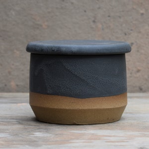 Peut inclure: Un beurrier en c&eacute;ramique fait &agrave; la main avec un couvercle. Le plat est cylindrique, avec une partie sup&eacute;rieure gris fonc&eacute; et une partie inf&eacute;rieure beige. Le couvercle est gris fonc&eacute; assorti. Le plat est pos&eacute; sur une surface en bois.