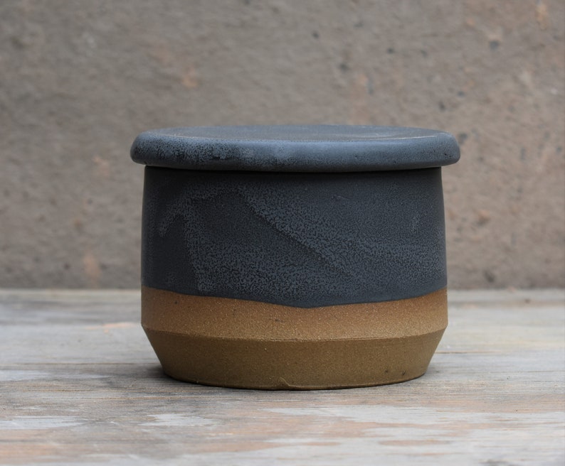 Peut inclure: Un beurrier en c&eacute;ramique fait &agrave; la main avec un couvercle. Le plat est cylindrique, avec une partie sup&eacute;rieure gris fonc&eacute; et une partie inf&eacute;rieure beige. Le couvercle est gris fonc&eacute; assorti. Le plat est pos&eacute; sur une surface en bois.