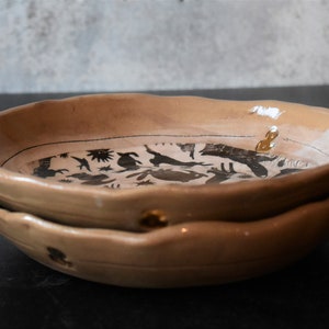 Puede incluir: Dos cuencos de cer&aacute;mica apilados con un esmalte marr&oacute;n claro. Los cuencos tienen un borde ondulado y est&aacute;n decorados con un dise&ntilde;o de silueta negra de animales y plantas.