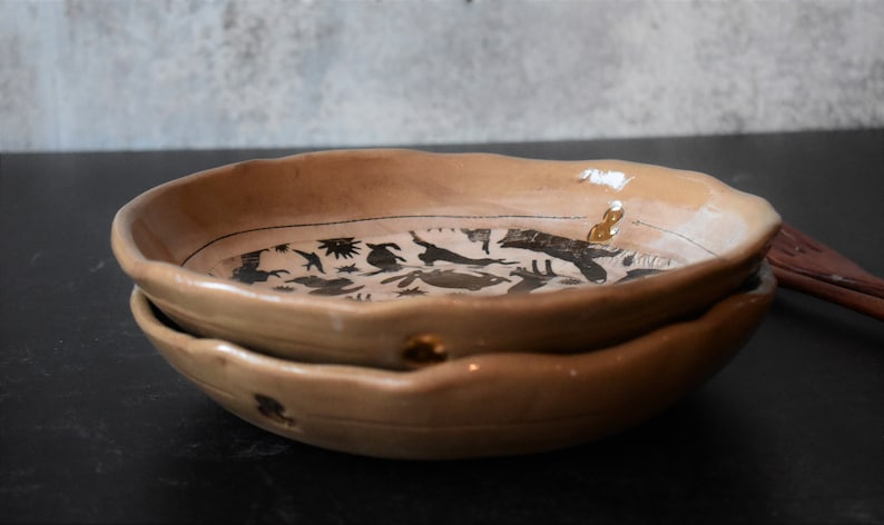 Puede incluir: Dos cuencos de cer&aacute;mica apilados con un esmalte marr&oacute;n claro. Los cuencos tienen un borde ondulado y est&aacute;n decorados con un dise&ntilde;o de silueta negra de animales y plantas.